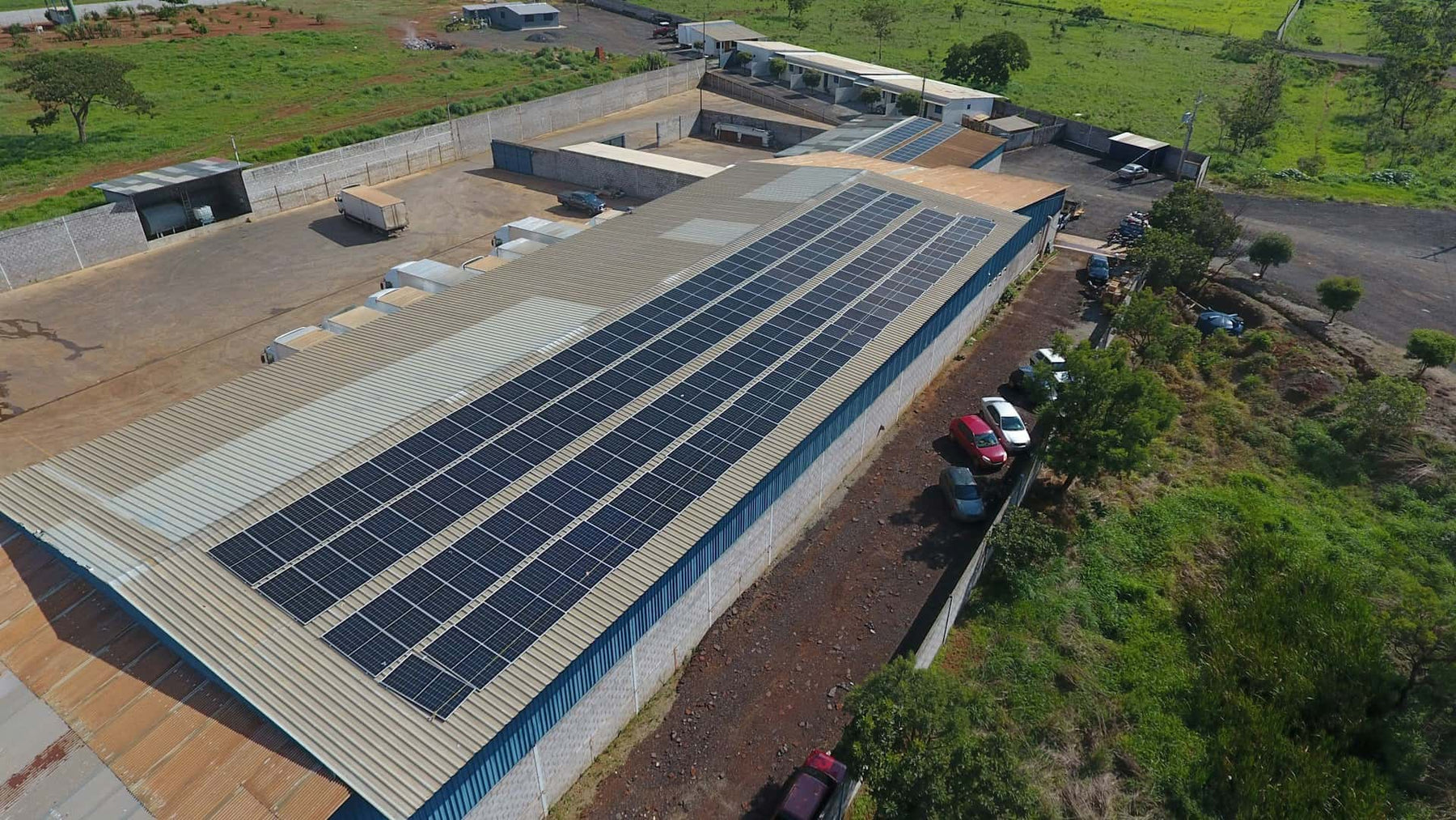Aerial view of an industrial premises with solar panels on its roof, surrounded by greenery and trucks.
